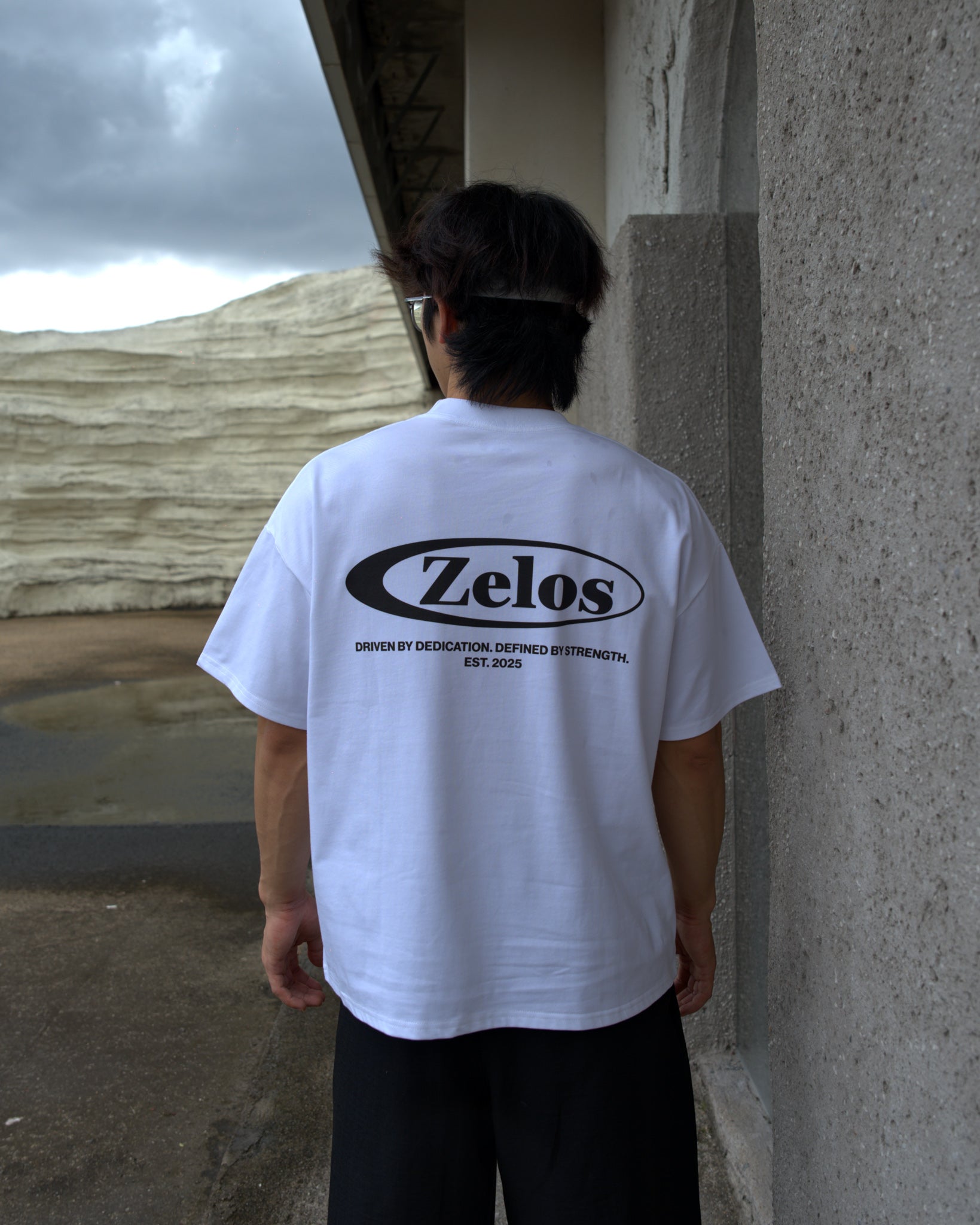 Male model wearing Zelos Tee White in size M, back view with Zelos logo print