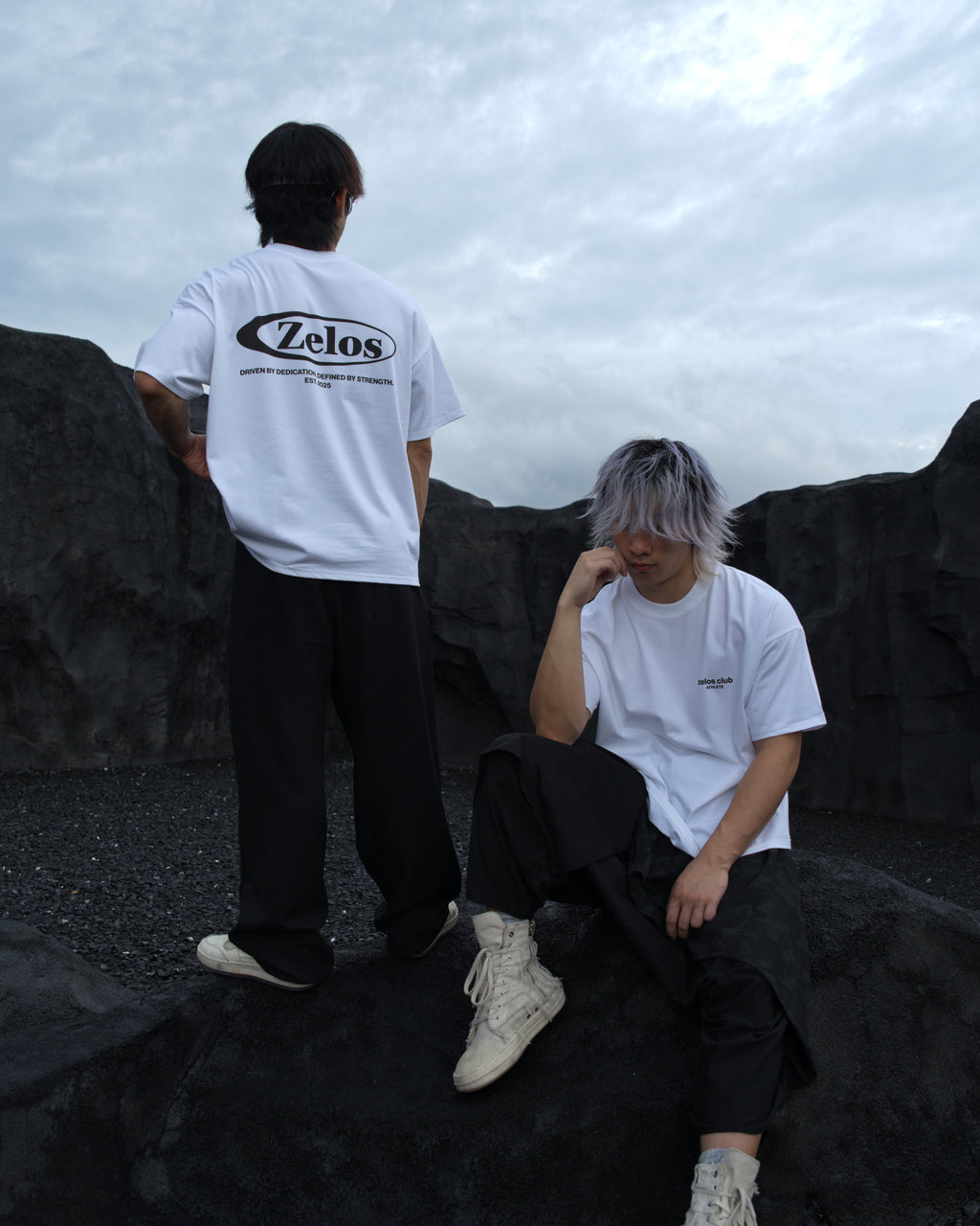 Two models wearing Zelos Tee White on volcanic rock, back and front views