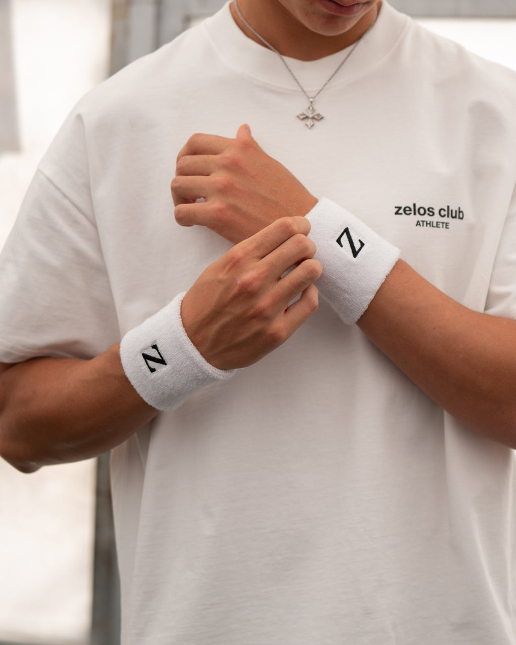A close-up of a person wearing a white athletic t-shirt and white wristbands with black logos. The t-shirt has 'zelos club ATHLETES' printed on the chest, and the wristbands feature a bold black 'Z' logo.